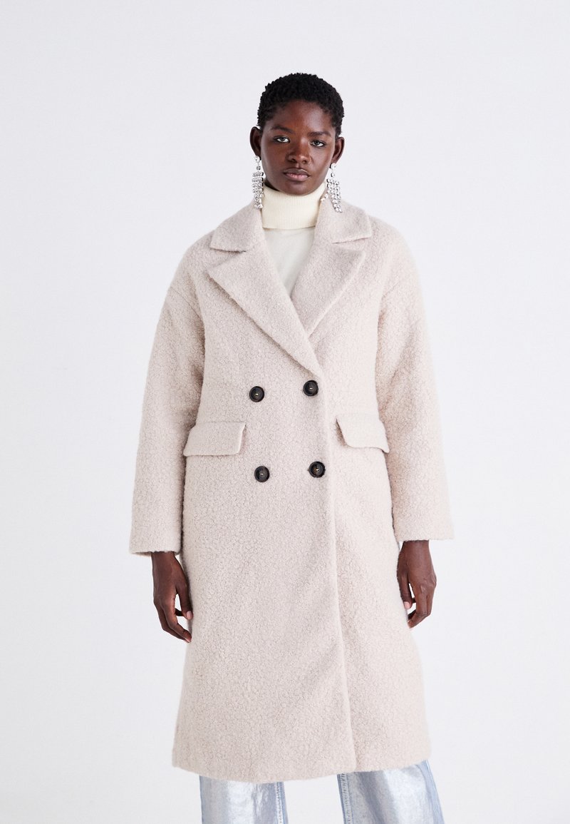 Cappotto di colore crema, doppiopetto, con finitura testurizzata, collo oversize, due tasche frontali e bottoni neri.
