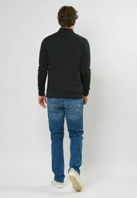 Svart langermet genser med høy krage, sammen med lyseblå jeans og hvite joggesko, sett fra baksiden.