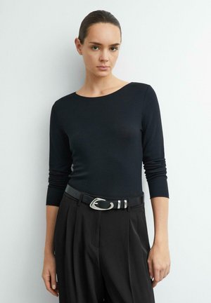 Frau mit dunkel zurückgebundenem Haar, die ein eng anliegendes schwarzes Langarmshirt und hoch taillierte schwarze Hose mit einem Gürtel mit silberner Schnalle trägt.
