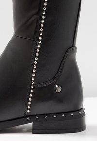 Stivaletto alla caviglia in pelle nera con dettaglio di perline argentate lungo la cucitura laterale e logo metallico sul tallone esterno. Suola in legno.