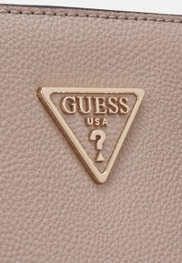 Cuir beige avec une surface texturée ; présente un logo triangulaire doré portant l'inscription "GUESS USA" et un symbole de point d'interrogation.