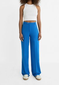 Pantalon large bleu en tissu lisse avec taille haute, doté d'une fermeture à boutons, de poches latérales et d'une coupe décontractée. Associé à un crop top blanc.