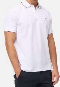 Polo-shirt blanc avec un col classique, des manches courtes, des accents rouge et marine sur le col et les poignets, avec un petit logo sur la poitrine gauche.