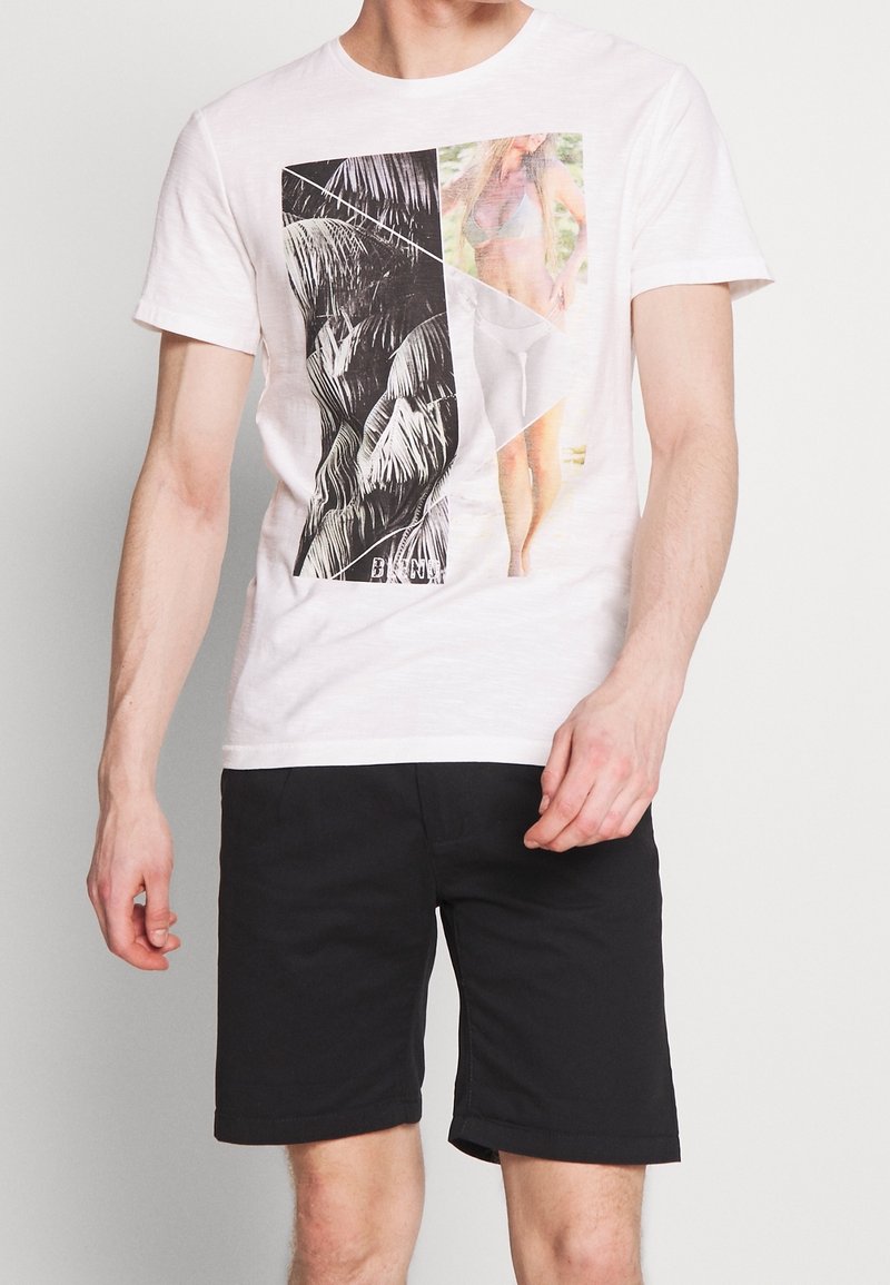 Homme portant un t-shirt blanc avec un collage photo en noir et couleur de feuilles de palmier et femme en bikini, assorti à un short noir.