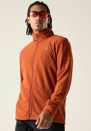Young man wearing an orange zip-up jacket and reflective red sport sunglasses against a plain light background.