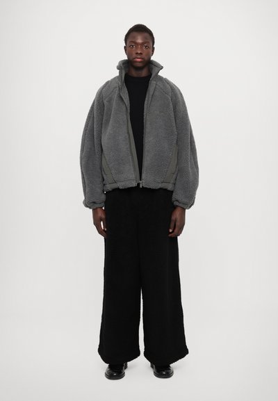 Jachetă gri din fleece cu guler înalt și închidere cu fermoar, asociată cu pantaloni largi din lână neagră. Țesături texturate și croială lejeră.