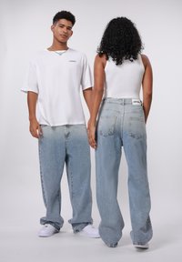 Afslappet hvid oversized t-shirt, blå højtaljede baggy jeans med ublegede områder, og hvide sneakers. Modellen har en tætsiddende hvid tanktop på.
