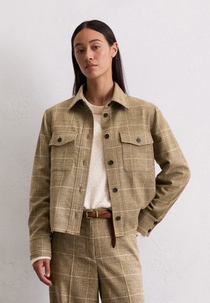 Vrouw gekleed in een beige geruite blazer en bijpassende broek met een bruine riem en een crèmekleurige gebreide trui, staande tegen een plain witte muur.