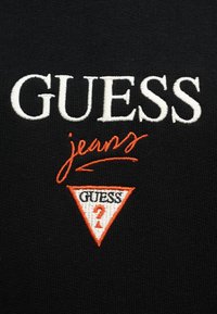 Το λογότυπο GUESS κεντημένο με λευκά γράμματα, με τη λέξη 'jeans' σε πορτοκαλί γραφή και ένα τρίγωνο με πορτοκαλί περίγραμμα που περιέχει τη λέξη 'GUESS' και ένα ερωτηματικό σε μαύρο ύφασμα.