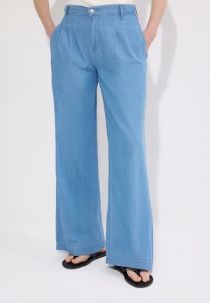 Lichtblauwe wijde broek, gedragen met zwarte teenslippers, handen in de zakken, vanaf de taille tot aan de voeten afgebeeld tegen een effen achtergrond.