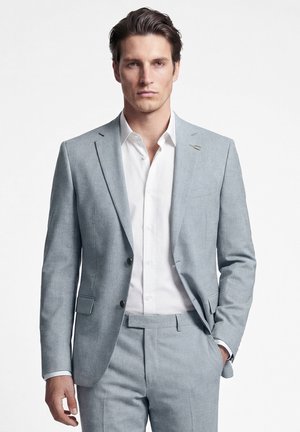 Man met donker haar, gekleed in een lichtgrijs pak en een wit overhemd, staande met één hand in de zak tegen een effen witte achtergrond.
