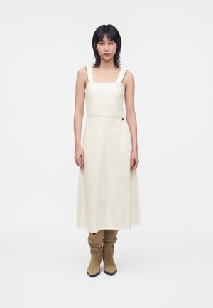 Femme aux cheveux noirs portant une robe midi crème sans manches et des bottes en daim camel montantes jusqu'au genou, debout devant un fond blanc uni.