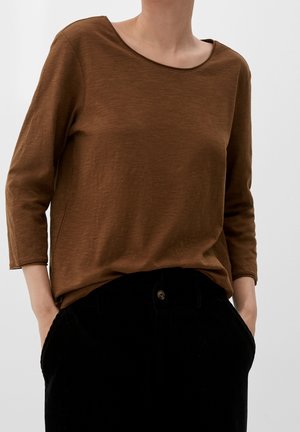 Persoon draagt een bruin shirt met lange mouwen, ingestopt in een hoog uitgesneden zwarte corduroy broek, met handen in de zakken, tegen een effen achtergrond.