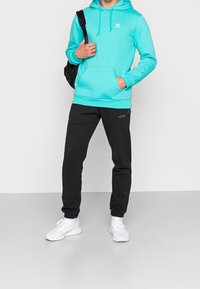 Tyrkysová mikina s kapucí, přední kapsou a šňůrkami, v kombinaci s černými tepláky s nášivkou loga. Celý outfit doplňují bílé tenisky.