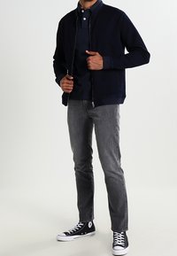 Námořnická bomber bunda na zip přes tmavé polo tričko, spojená se šedými jeansy a černými vysokými teniskami. Hladká tkanina, přiléhavý střih.