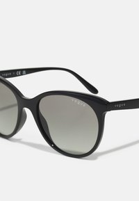 Ochelari de soare din plastic negru cu lentile gri degradé, formă rotundă de cat-eye și accente argintii pe braț, având logo-ul "vogue".