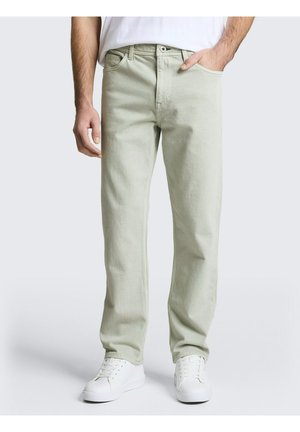 Man die lichtbeige rechte jeans en witte sneakers draagt, met één hand in de zak staand tegen een effen achtergrond.