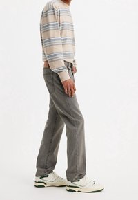 Persona vestida con una camisa de rayas beige y azul de manga larga, jeans grises de pierna recta y zapatillas blancas con acentos verdes, de pie de lado.