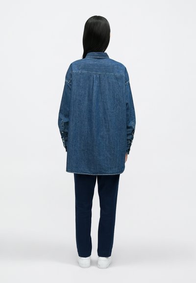 Τζιν πουκάμισο με άνετ�η εφαρμογή, μακριά μανίκια και μανσέτες με κουμπιά· συνδυασμένο με ναυτικές μπλε παντελόνες και λευκά αθλητικά παπούτσια.