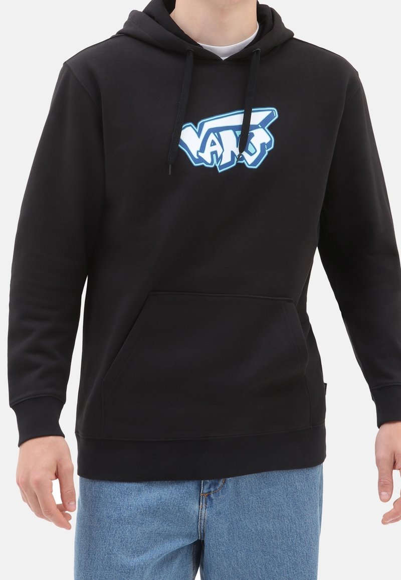 Homme portant un sweat à capuche noir Vans avec un logo bleu et blanc, associé à un jean bleu clair, sur fond blanc.