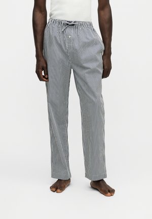 Homme portant un pantalon de pyjama rayé vertical noir et blanc avec une taille à cordon, debout pieds nus sur un fond blanc uni.