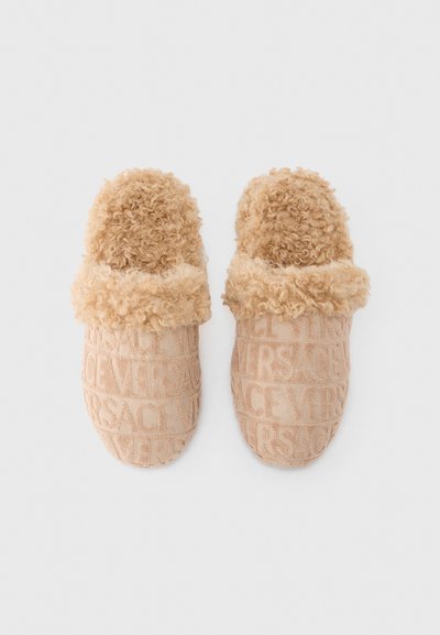 Chaussons beige en tissu texturé, avec une bande supérieure duveteuse. Logo "VERSACE" discrètement embossé sur tout le corps. Design confortable.