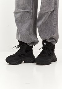Zapatillas negras de caña alta con tejido texturizado, cuello acolchado, suela voluminosa y diseño con cordones. Combinadas con pantalones deportivos gris sueltos.