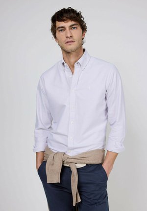 Joven con camisa de botones color púrpura claro, suéter beige atado alrededor de la cintura y pantalones azul oscuro, de pie frente a un fondo liso.