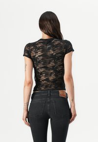 Μαύρη μπλούζα από δαντέλα με κοντά μανίκια, εφαρμοστή γραμμή και ημι-διαφανή υφή, σε συνδυασμό με σκούρα τζιν που έχουν ετικέτα στο πίσω μέρος.