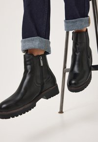 Botas de cuero negras hasta el tobillo con cremallera lateral, suela resistente y acentos cosidos, combinadas con jeans de mezclilla oscura enrollados en los puños.