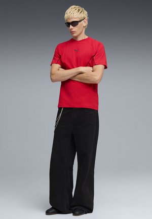 Maglietta sportiva rossa con accenti neri e logo del marchio, abbinata a pantaloni neri ampi con una catena decorativa. Il modello indossa occhiali da sole.