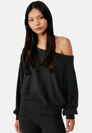 Jonge vrouw met lang donker haar, gekleed in een donkergrijze oversized trui met blote schouders en bijpassende broek, staand tegen een effen achtergrond.