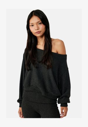 Jonge vrouw met lang donker haar, gekleed in een donkergrijze oversized trui met blote schouders en bijpassende broek, staand tegen een effen achtergrond.