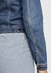 Veste en denim bleu clair, dotée d'une fermeture à boutons avec des accents foncés, associée à un pantalon rayé bleu et blanc, mettant en avant un détail de poche.