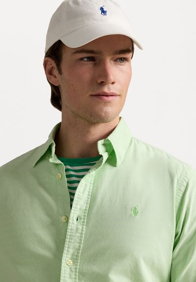 Camisa ligera de color verde con botones y un pequeño logotipo bordado, superpuesta sobre una camiseta a rayas verdes y blancas; gorra blanca de béisbol con logotipo.