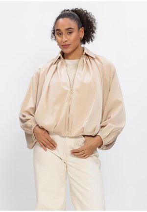 Vrouw met krullend haar, gekleed in een beige jas met rits en crème broek, staand met handen in de zij tegen een effen achtergrond.