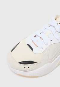 Deportiva con parte superior de malla color crema, acentos en negro y cordones blancos. Cuenta con una suela de goma texturizada y detalles de costura sutiles.