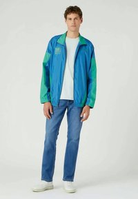 Sininen ja vihreä kevyt takki korkean kauluksen, elastisten hihojen ja vetoketjullisen etuosan kanssa. Yhdistettynä vaaleansinisiin farkkuihin ja valkoisiin lenkkareihin.