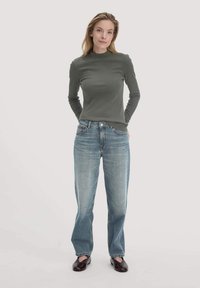 Langärmliges, olivgrünes Oberteil mit hohem Ausschnitt, kombiniert mit hellblauen, geraden Jeans und dunkelbraunen Slippern.