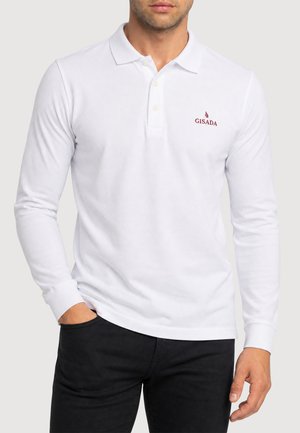 Mann trägt weißes Langarm-Poloshirt mit Kragen und zwei Knöpfen, mit kleinem roten "GISADA"-Logo auf der linken Brust, kombiniert mit schwarzen Hosen.