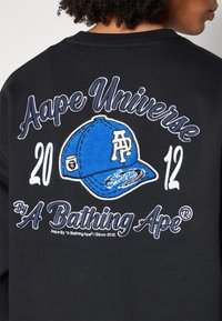 Dos de sweat-shirt noir avec un motif de casquette bleue brodée et un texte blanc « Aape Universe 2012 By A Bathing Ape » autour.