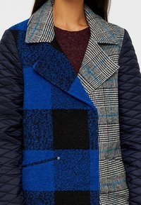 Donna che indossa un cappotto in patchwork con davanti in lana check blu e nera, colletto a quadri bianco e nero, e maniche in tessuto trapuntato color navy.