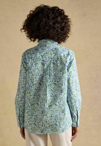 Vrouw met krullend haar die een blauwgroen bloemenprint shirt met lange mouwen draagt, met haar rug naar de camera toe staand tegen een beige achtergrond.