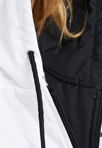 Närbild av en vit jacka med svart dragsko och dragkedja, bärs över ett svart plagg, med strån av ljusbrunt hår synliga.