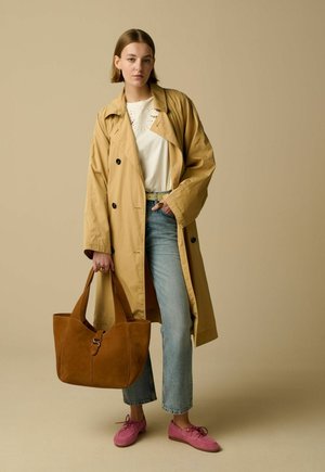 Vrouw draagt beige trenchcoat, wit overhemd, blauwe jeans, roze schoenen, houdt een grote bruine leren draagtas vast, staand tegen een beige achtergrond.