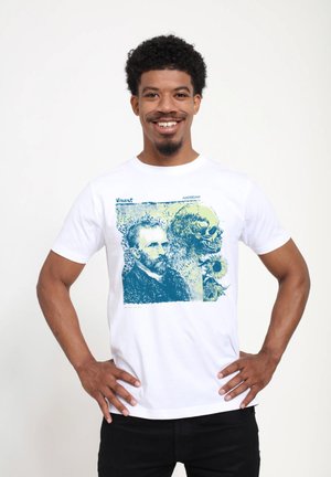 Usmívající se muž s kudrnatými vlasy stojí s rukama na bocích, oblečený v bílém tričku s designem Vincenta van Gogha, lebky a slunečnic.