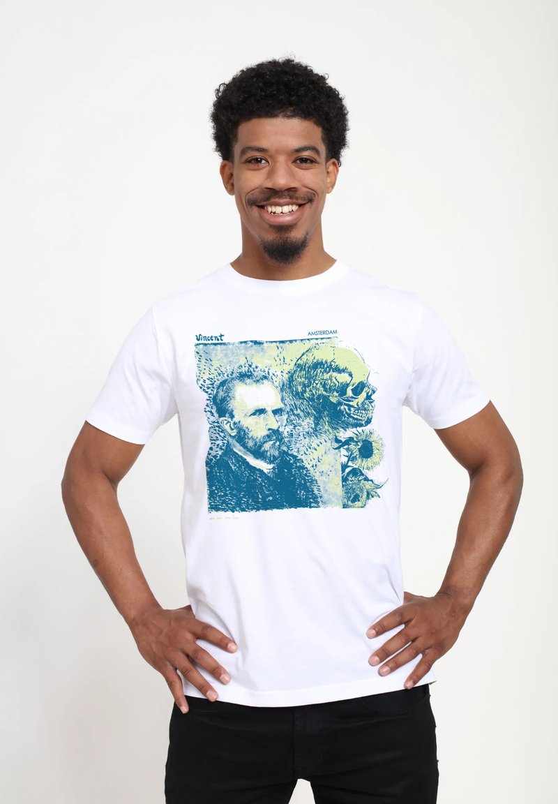 Usmívající se muž s kudrnatými vlasy stojí s rukama na bocích, oblečený v bílém tričku s designem Vincenta van Gogha, lebky a slunečnic.