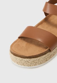 Sandalo in pelle marrone con due ampie cinghie, soletta simile alla suede e suola in iuta intrecciata. Design piatto senza hardware visibile o motivi.