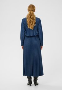 Hosszú, pliszírozott sötétkék ruha gombos hátrésszel; puha anyagból készült, hosszú ujjú és a dereka szoros, fekete csizmával párosítva.