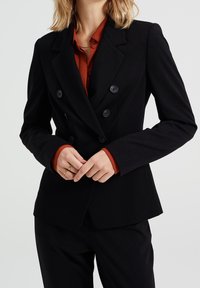 Svart dubbelknäppt blazer med två rader knappar, skräddarsydd passform, med slagkrage. Bärs över en mörkröd skjorta. Slät tygtextur.